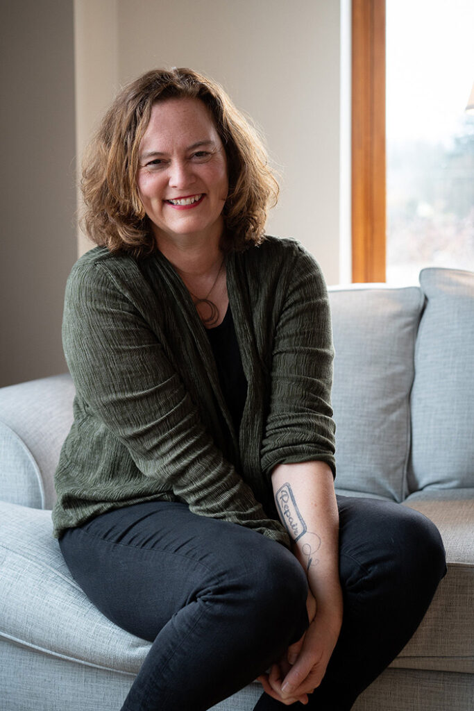 Sarah Murphy Kangas, wearing a green sweater and black pants, sits on a grey couch. 